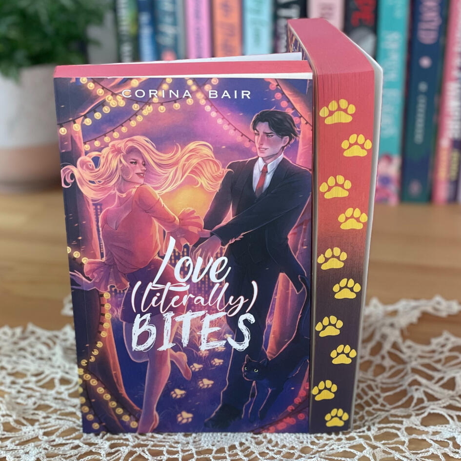 The cover of Love (Literally) Bites: A paranormal romcom by Corina Bair featuring a woman with blonde hair reaching back for a dark-haired man in a suit as she trails pawprints instead of footprints. There's a black cat leaping after them and a city in the